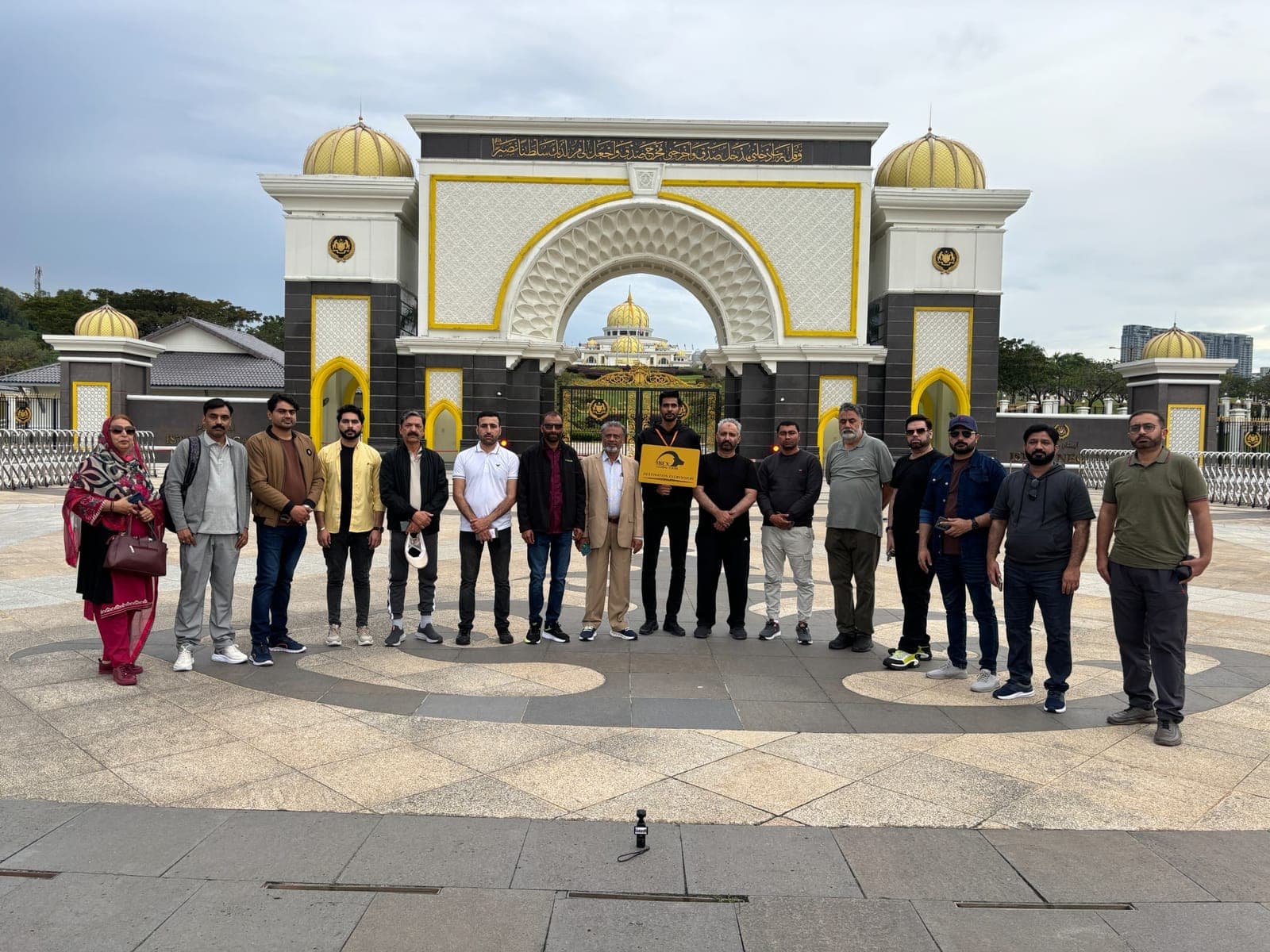 An IBEX group photographed in front of the gates of the Istana Negara in Kuala Lumpur, one traveller holding an IBEX Global Tours board